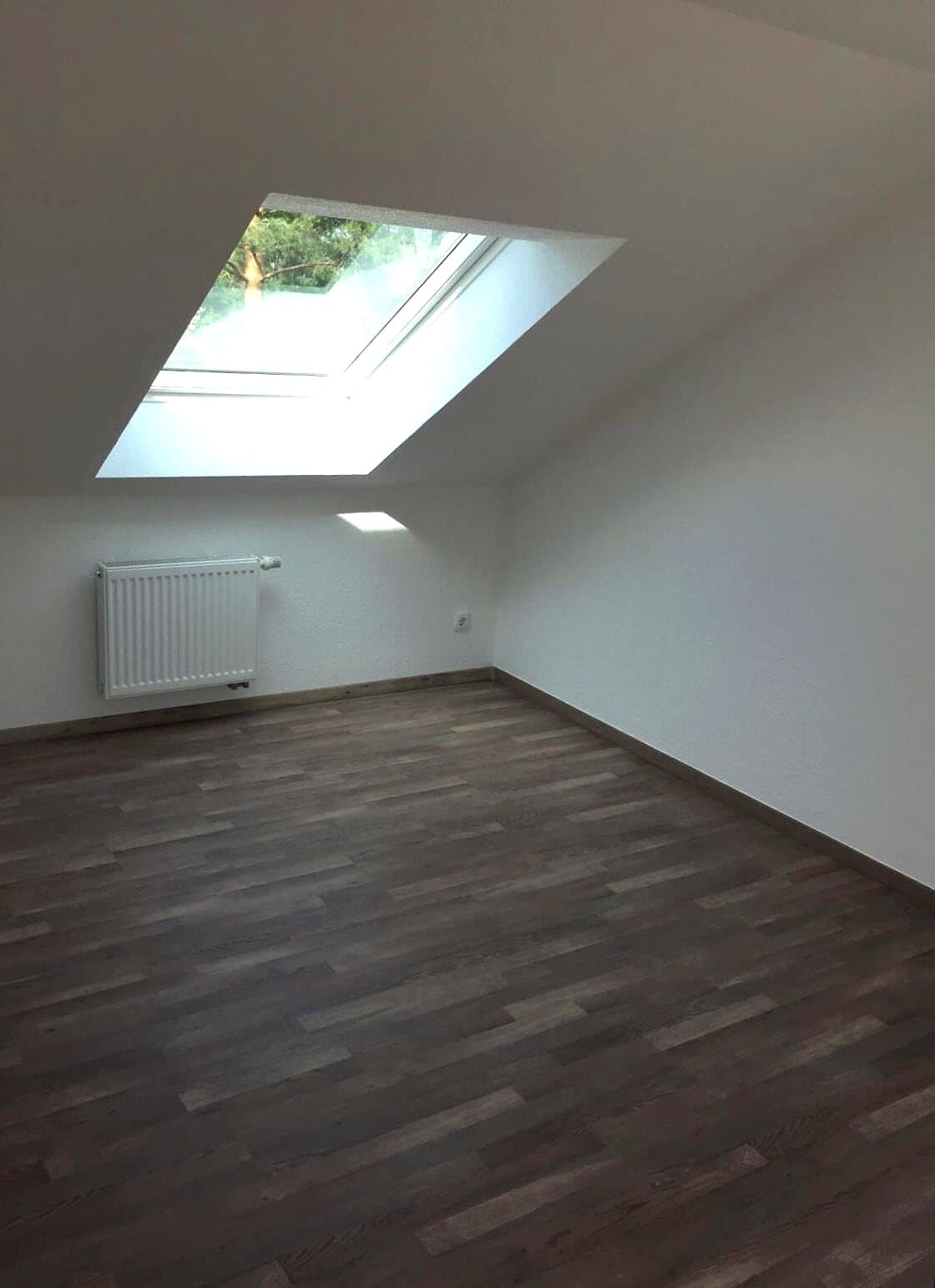 Fertiges Dachzimmer mit Dachfenster, weißen Wänden und dunklem Dielenboden