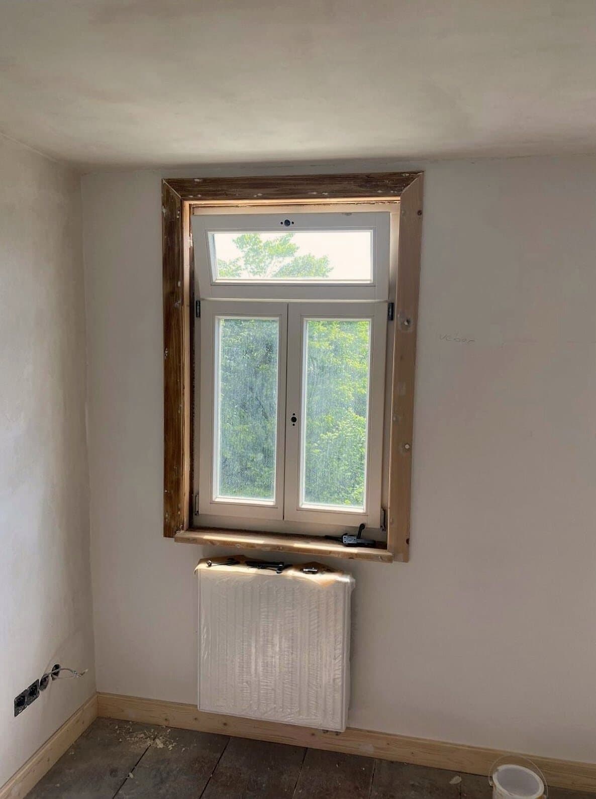 Frisch verputzte helle Wand mit Holzfenster, darunter ein mit Folie abgedeckter Heizkörper